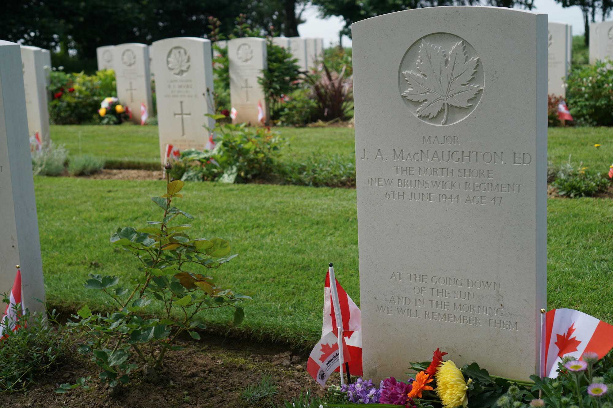 Colour photograph. Archie's grave, in Beny-sur-Mer Canadian War Cemetery. Flowers and small Canadian flags are planted in the ground at its base.